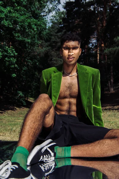 African american man in green blazer and black shorts sitting with closed eyes in park on reflective surface - foto de stock