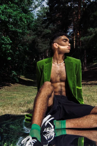 African american man with closed eyes sitting in green blazer on mirror in forest - foto de stock