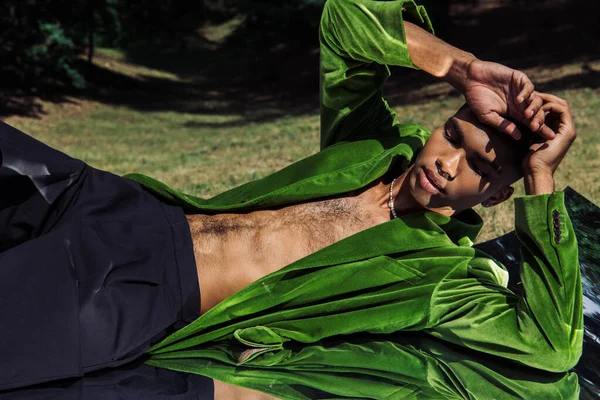 Young african american man in trendy green blazer lying with hands near head on reflective surface in park — Stockfoto