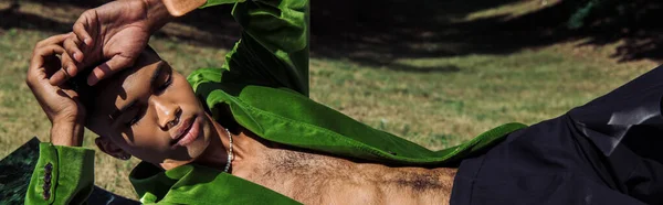 Fashionable african american man in green velvet blazer lying on lawn with hands near head, banner — Foto stock