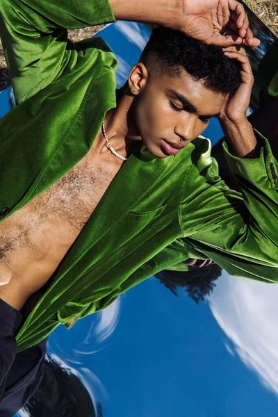 Top view of young african american man in green velvet blazer lying on mirror outdoors — стоковое фото