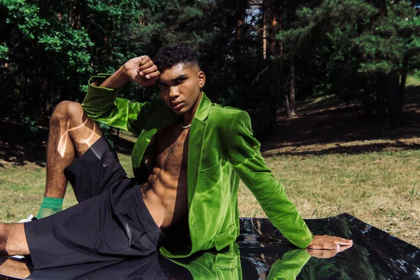 Trendy african american man in green blazer and black short sitting on mirror in park and looking at camera — стоковое фото