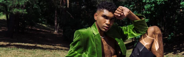 Trendy african american man in green velvet blazer looking at camera while sitting in park, banner — стоковое фото