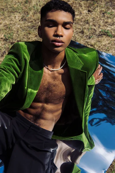 Young african american man in beads and velvet blazed sitting on lawn and reflective surface — Stockfoto