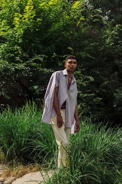 African american man in trendy shirt and white trousers looking at camera in green park — стоковое фото