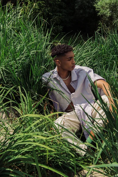 Модний афроамериканський чоловік сидить на зеленій траві і дивиться в сторону — стокове фото