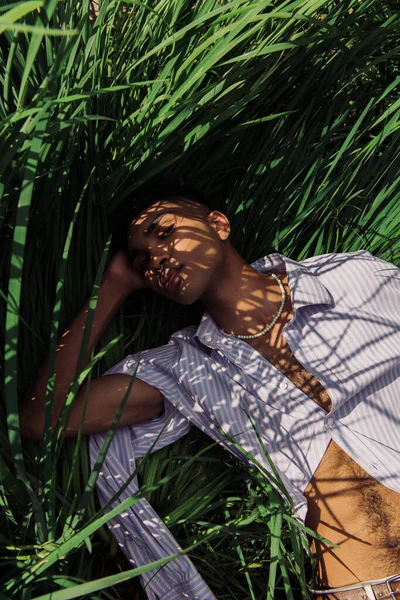 Top view of african american man in blue striped shirt lying on green grass — Foto stock