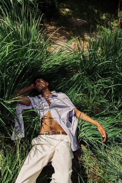 Високий кут зору афроамериканця в стильному літньому вбранні спить на зеленій траві — стокове фото