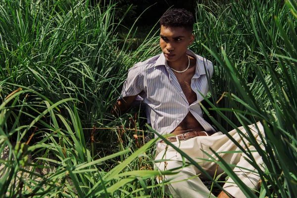 Молодий афроамериканський чоловік у блакитній сорочці дивиться далеко, сидячи на траві в парку — стокове фото