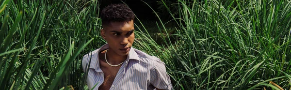 Stylish african american man looking away while sitting on green grass, banner — стоковое фото