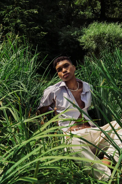 Fashionable african american man sitting on green grass and looking at camera — Fotografia de Stock