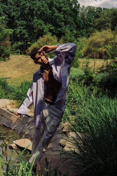 Stylish african american man with hand near face posing near pond in park — Photo de stock