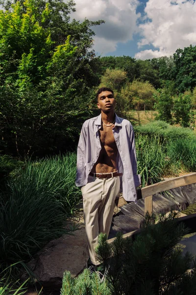 African american man in blue shirt and white trousers posing near lake in park — Photo de stock
