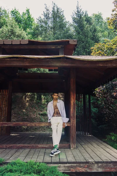 Full length of african american man in trendy summer outfit standing in wooden patio in park - foto de stock