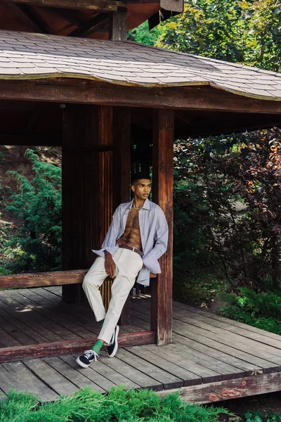 African american man in white trousers and blue shirt posing on wooden terrace in park — Photo de stock