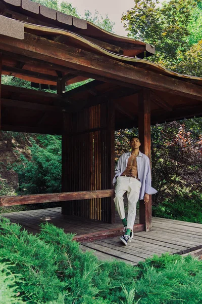 Young african american man in stylish clothes posing in wooden gazebo in green park - foto de stock