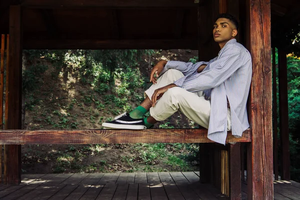 African american man in blue striped shirt and sneakers sitting in wooden patio — стоковое фото