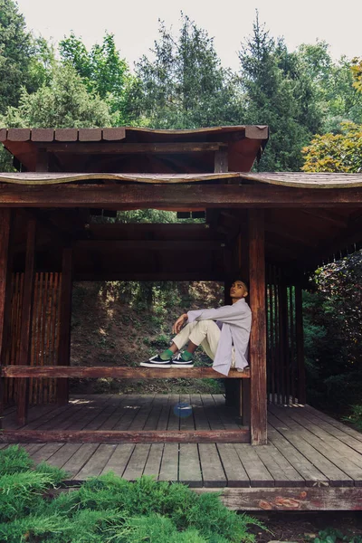 Full length of african american man in summer clothes and sneakers sitting in wooden gazebo - foto de stock