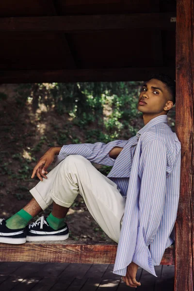 Young african american man in blue and striped shirt sitting on wooden terrace in park — Foto stock