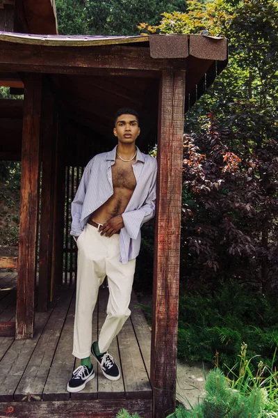 Full length of african american man in stylish clothes looking at camera in wooden gazebo — Photo de stock