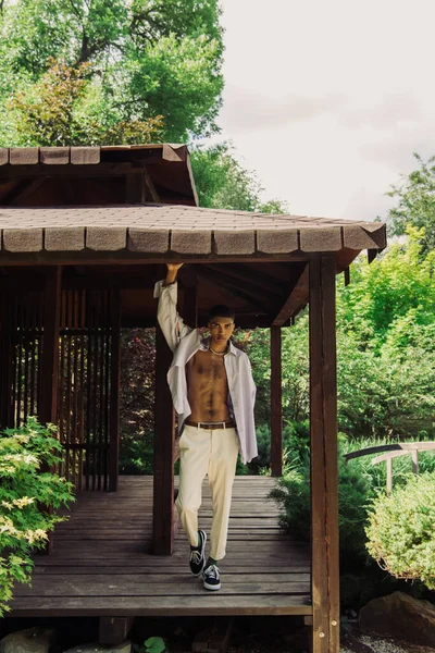 Full length of african american man in trendy clothes posing in wooden patio in summer park - foto de stock