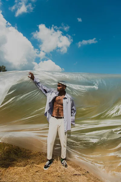 African american man in blue shirt and white trousers near polyethylene film in field — Fotografia de Stock