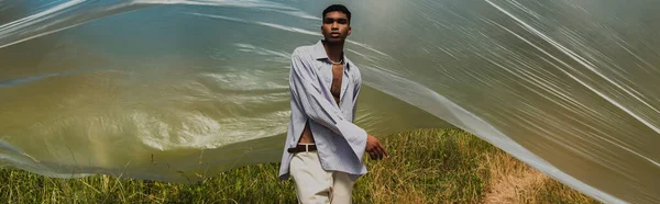 Young african american man in trendy summer outfit near cellophane film in field, banner — Foto stock