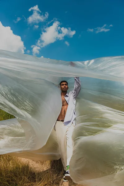 Young african american man in trendy clothes standing with closed eyes under polyethylene and cloudy sky — стоковое фото