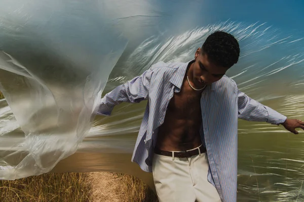 Young african american man in shirt standing under cellophane in field — Fotografia de Stock