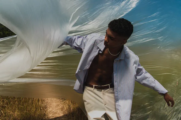 Young african american man in shirt and trousers posing under polyethylene outdoors — стоковое фото