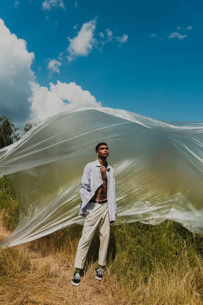 Fashionable african american man with closed eyes under polyethylene film in grassy meadow — Foto stock