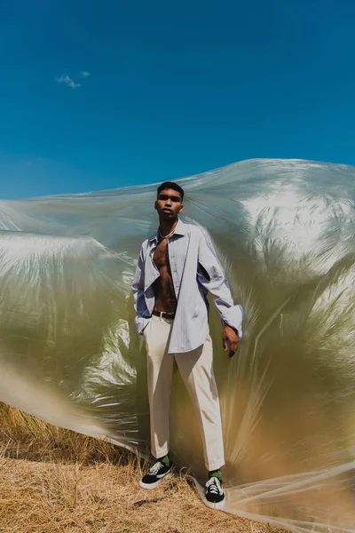 Full length of african american man in summer outfit standing in meadow near polyethylene film — Fotografia de Stock