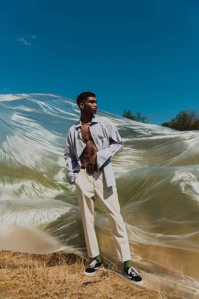 Молодий афроамериканський чоловік з рукою в кишені білих штанів, що стоять на лузі біля поліетиленової плівки — стокове фото