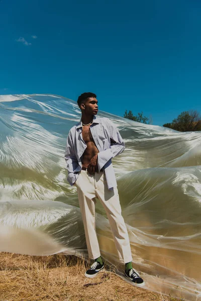 Young african american man with hand in pocket of white trousers standing in meadow near polyethylene film — Photo de stock