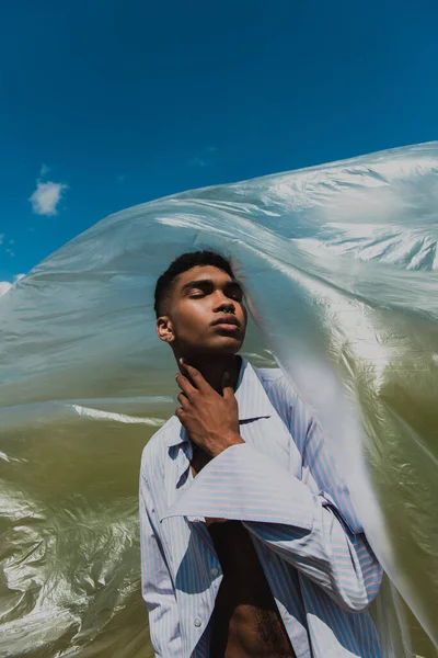 Stylish african american man with closed eyes touching neck while posing near polyethylene under blue sky — Fotografia de Stock