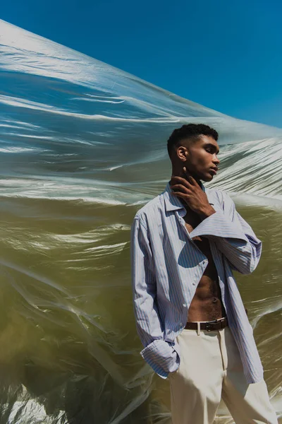 African american man in trendy shirt touching neck near cellophane film outdoors — стоковое фото