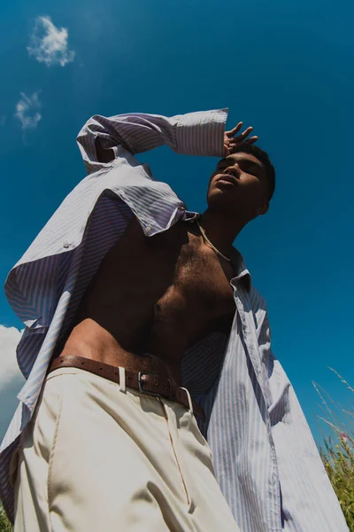 Low angle view of african american man in blue striped shirt posing with closed eyes against blue sky — Foto stock