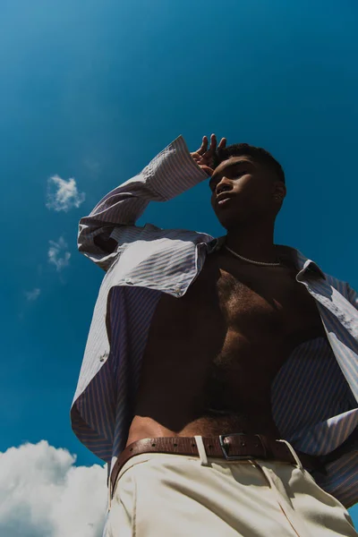 Low angle view of african american man in striped shirt posing with hand near head against blue sky — Photo de stock