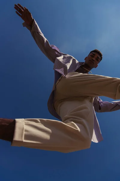 Bottom view of african american man in white pants jumping under blue sky — стоковое фото