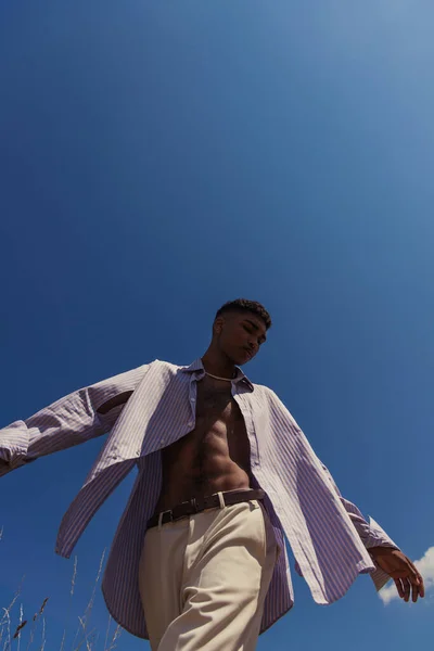 Low angle view of african american man in striped shirt and white trousers against cloudless sky - foto de stock