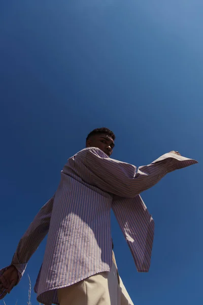 Low anlge view of african american man in striped shirt looking at camera under blue cloudless sky — Photo de stock