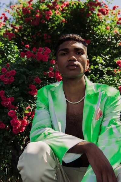 African american man in beads and trendy blazer looking at camera near blossoming bushes — Fotografia de Stock