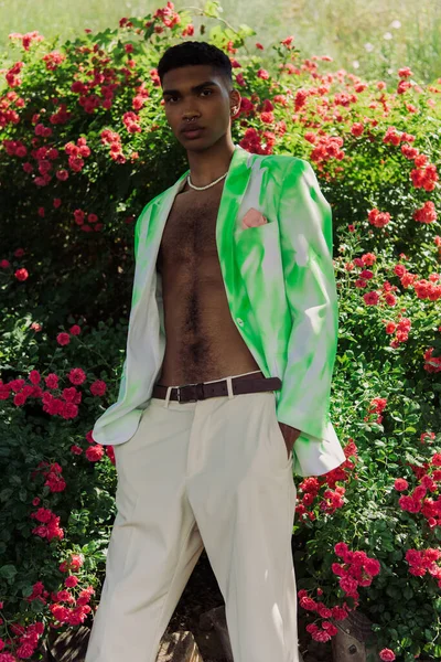 Young and stylish african american man standing with hands in pockets near blossoming bushes — Fotografia de Stock