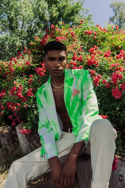 African american man in stylish blazer and beads sitting near blossoming bushes and looking at camera — Stock Photo