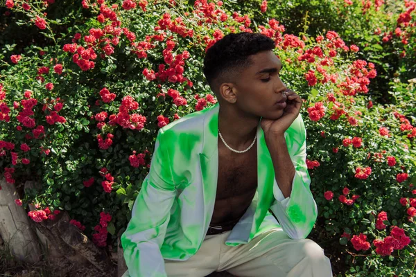 Young african american man in trendy blazer sitting near blossoming bushes in park — Photo de stock