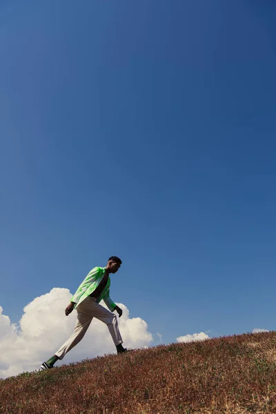 Side view of african american man walking in grassy field under blue sky with white clouds - foto de stock