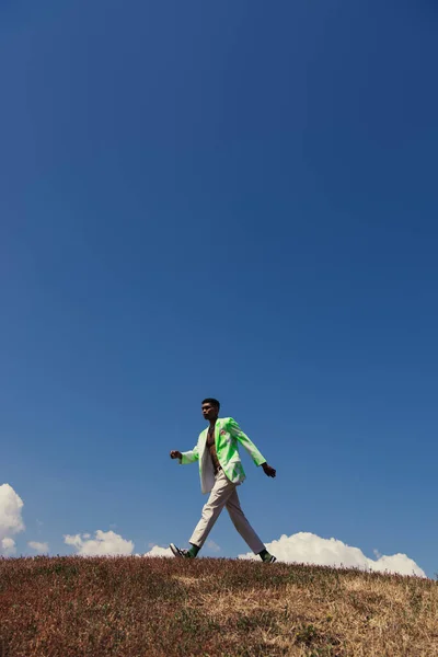 Side view of african american man in blazer and pants walking in field under clear sky — Fotografia de Stock