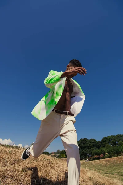 Молодий афроамериканський чоловік в білому і зеленому блістері біжить під блакитним небом на лузі — стокове фото