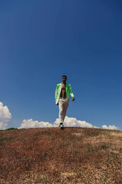 Full length of african american man in trousers and blazer walking in field under blue sky with white clouds — Fotografia de Stock