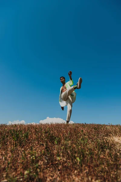 Trendy african american man posing on one leg in grassy field under blue sky — Stockfoto